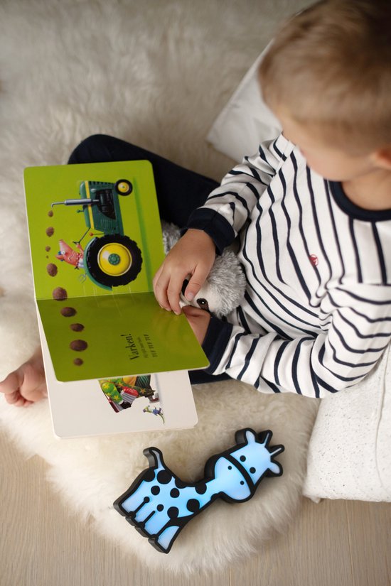 Blauw oplichtend giraf-nachtlampje naast een jongen die een boek leest op een wit vloerkleed.
