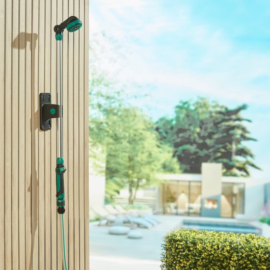 Een zilveren sproeilans met groene accenten hangt aan een muur van houten planken, met een tuin en terras op de achtergrond.