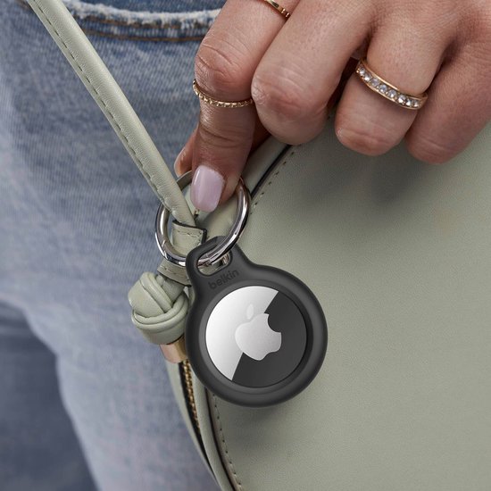 Hand met ringen houdt een lichte tas vast met een zwarte Belkin AirTag houder met sleutelhanger.