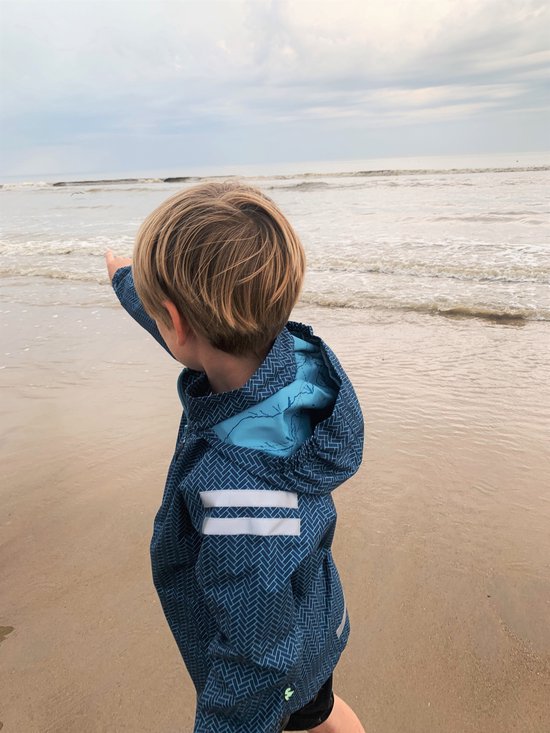 Jongen met blond haar wijst naar de zee, draagt een blauw jack met visgraatpatroon.