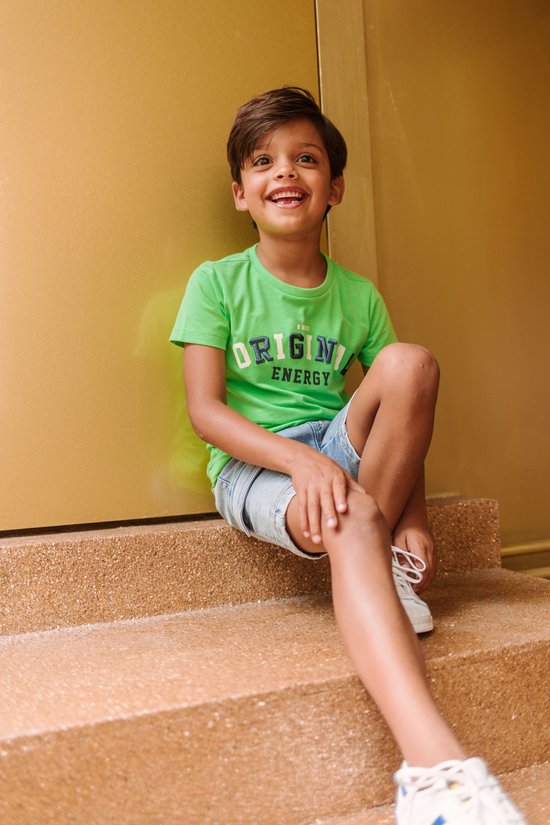 Jongetje met donker haar, lachend op de trap, met een felgroen T-shirt met 'ORIGINAL ENERGY' en een spijkerbroek.