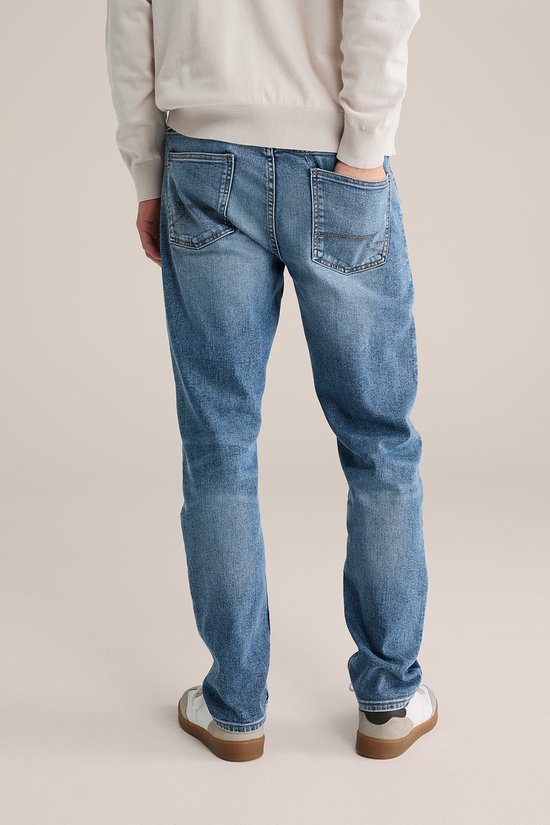 Achterkant van een man die een beige trui en een blauwe jeans met lichte wassing draagt.
