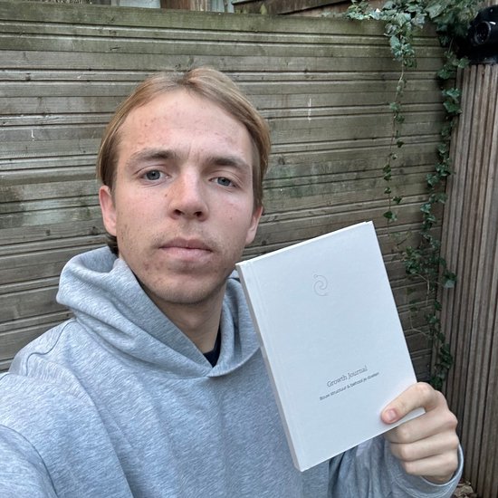 Jonge man in grijze hoodie houdt een wit notitieboekje vast met de tekst 'Growth Journal'.