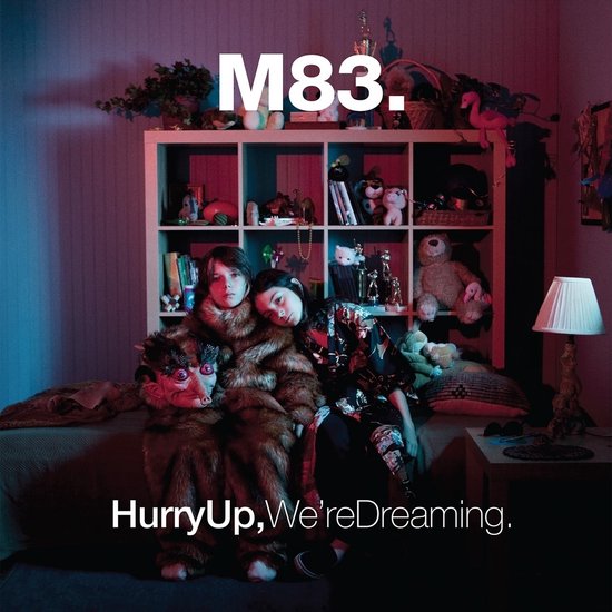 Twee personen zittend op een bed, gekleed in bontjassen, met de tekst "M83. Hurry Up, We're Dreaming."