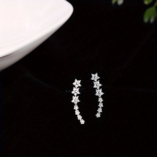 Zilverkleurige oorclimbers met witte zirkonia bloemetjes, liggend op een zwarte achtergrond.