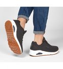 Voeten in zwarte Skechers sneakers met witte zolen, zittend op de grond.