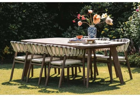 Een rechthoekige houten tuintafel met zes bijpassende stoelen, omringd door groen. De stoelen hebben een gevlochten zitting en rugleuning in lichte en donkere tinten.