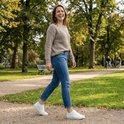 Lachende vrouw in een park, draagt een beige trui, blauwe jeans en witte sneakersokken.