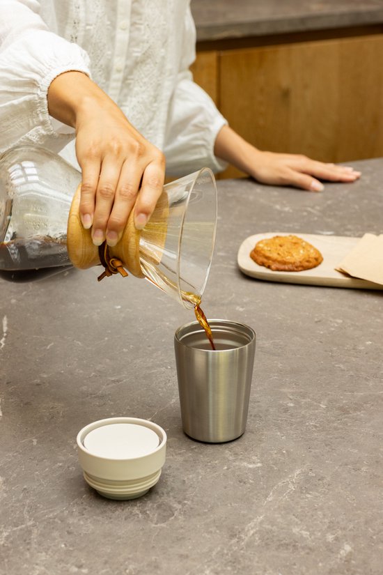 Hand schenkt koffie uit een glazen koffiezetapparaat in een zilverkleurige thermosbeker.