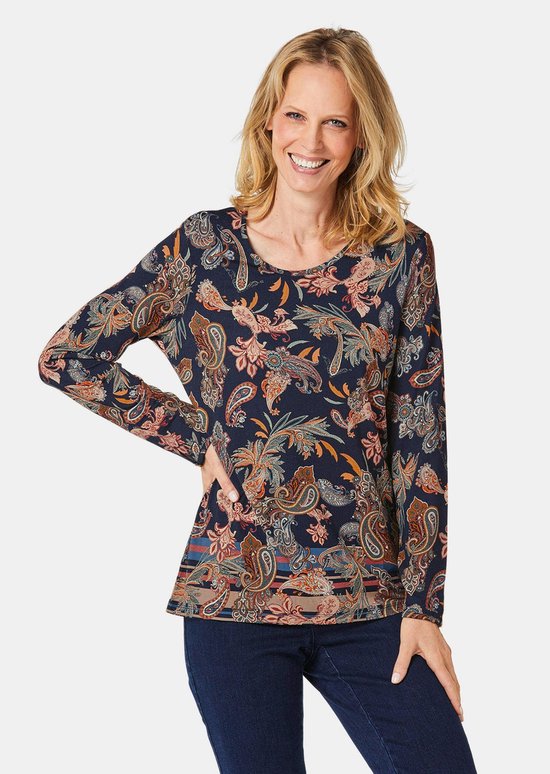 Femme aux cheveux blonds souriante, portant un haut bleu marine à imprimé cachemire, main sur la hanche.