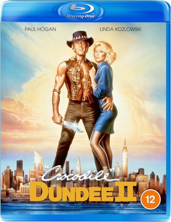 Blu-ray hoes van Crocodile Dundee II. Paul Hogan in cowboykleding met mes, Linda Kozlowski in blauwe jurk, met stadsgezicht.