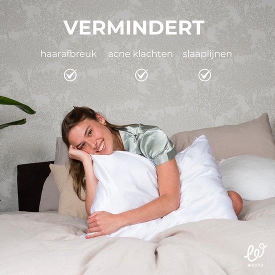 Een glimlachende vrouw in een satijnen pyjama ligt op bed met haar hoofd op een witte zijden kussensloop. De tekst zegt 'VERMINDERT haarafbreuk, acne klachten, slaaplijnen'.