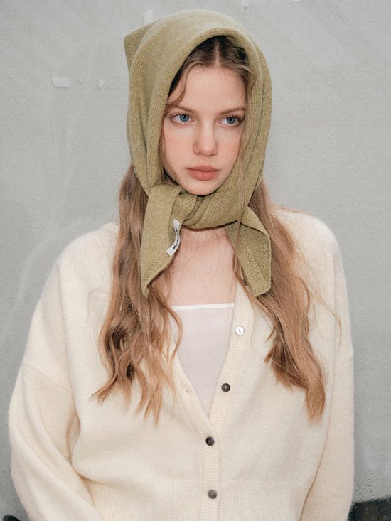 Model draagt een gebreide gebroken witte vest. De olijfgroene sjaal wordt als hoofddoek gedragen met de uiteinden geknoopt.