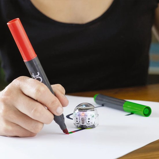 Persoon met zwarte trui tekent met een rode Ozobot marker op wit papier. Een kleine, transparante Ozobot robot staat naast de getrokken lijn. Een groene marker ligt op de achtergrond.