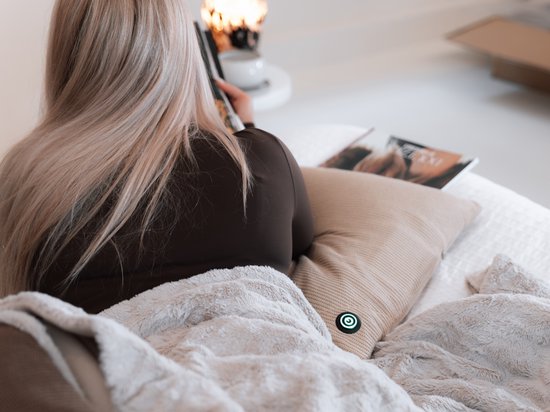 Vrouw met blond haar ligt onder een deken op een bank, met een beige kussen met groen logo.