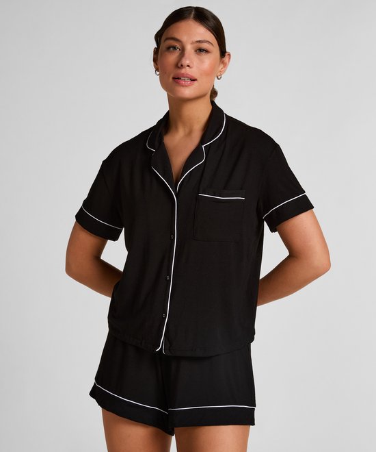 Model draagt een zwart pyjamashirt met korte mouwen en witte piping. Het shirt heeft een open kraag en een borstzakje.