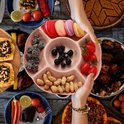 Roze snackbord gevuld met macrons, bessen, kerstomaatjes en pinda's, vastgehouden boven een tafel.