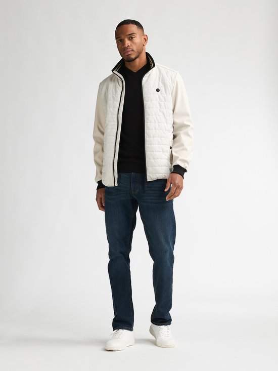 Man draagt een lichte beige gewatteerde jas met jeans en zwart T-shirt. De jas heeft beige mouwen en zwarte boorden.