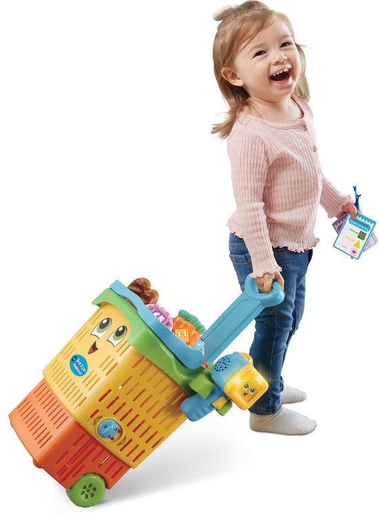 Blond meisje in roze trui, trekt een felgekleurde winkelwagen met speelgoedboodschappen.