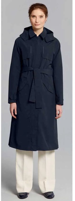 Vrouw in lange donkerblauwe jas met capuchon, ceintuur en witte broek, van voren gezien.