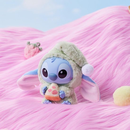 Een mintgroene Stitch pluche speelgoedje in slaapmutsje, met een pizzapunt in de handen, op roze heuvels.