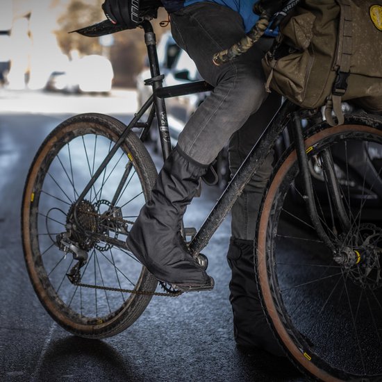 Een persoon op een fiets op een natte straat, draagt zwarte fietsoverschoenen.