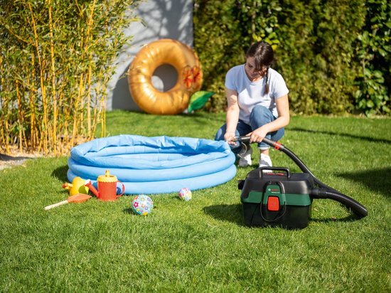 Vrouw in blauw T-shirt en spijkerbroek stofzuigt gras naast opblaasbaar zwembad. Ketelstofzuiger met groene en rode accenten.