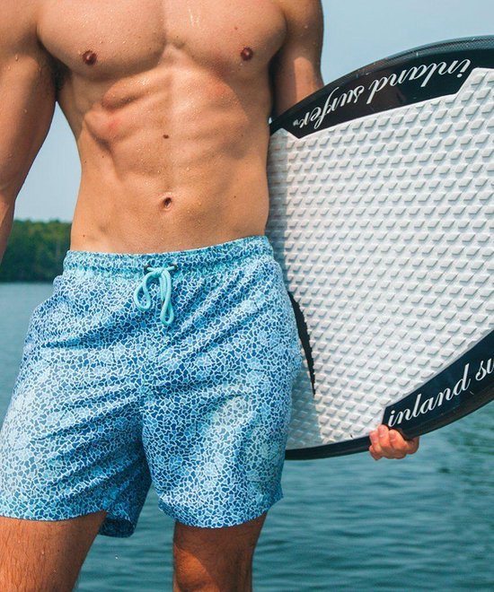 Gedetailleerde close-up van een gespierde man die een blauwe zwembroek draagt met een mozaïekprint en een surfplank vasthoudt.