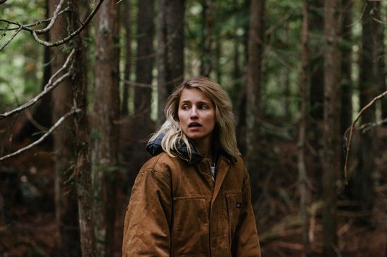 Blondine met oranje jas in bos, kijkt angstig weg.