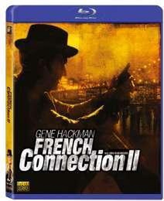 Gene Hackman in "French Connection II" Blu-ray hoes, met silhouet van man in hoed, vuurwapen en stadslichten.