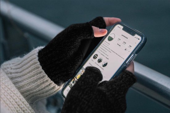 Vrouw met blonde haren en witte trui, draagt zwarte vingerloze handschoenen en houdt een telefoon vast.