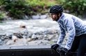 Man op een racefiets bij een rivier en weg, draagt een witte, doorschijnende fietsregenjas en een helm.