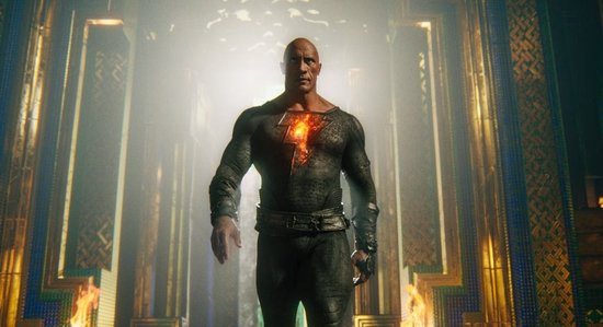 Dwayne Johnson in een zwart superheldenpak, met gouden bliksem op de borst, staat tussen pilaren met vlammen.