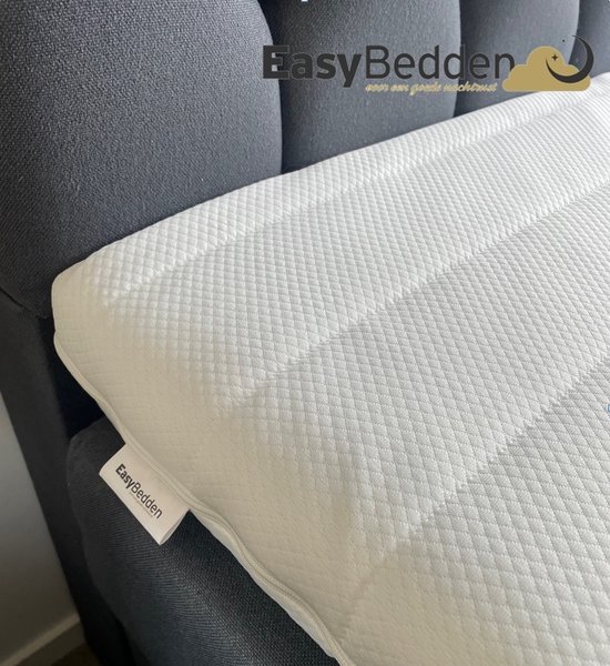 Witte topper met ruitpatroon, met een Easy Bedden label. Geplaatst op een bed met donkergrijze hoofdbord.