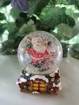 Prachtige sneeuwbol Kerstman met kerstbellen en cadeauzak leunend tegen schouw op sokkel in de vorm van een dak 5,5x8cm