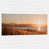 WallClassics - Muursticker - Zon Verdwijnend achter Bergen bij Strand - 100x50 cm Foto op Muursticker