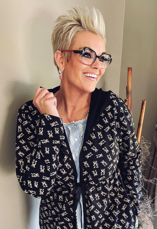 Une femme aux cheveux blonds et portant des lunettes, porte une chemise de nuit et un peignoir noir à imprimé ours en peluche.