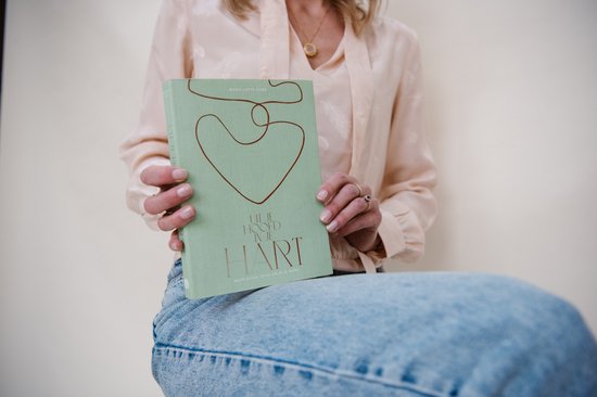 Detail van een vrouw in een lichtroze blouse en spijkerbroek die een groen boek vasthoudt met de tekst 'UIT JE HOOFD IN JE HART'.