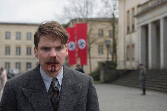 Een man met bloed uit zijn neus en mond, draagt een grijs pak, nazivlaggen op de achtergrond.