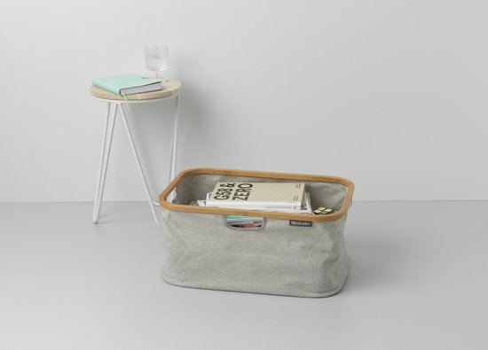 Grijze Brabantia wasmand met bamboe frame, gevuld met tijdschriften en een glas water op een bijzettafel.