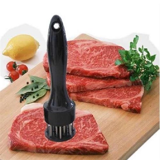 Attendrisseur à viande noir sur une planche en bois avec plusieurs steaks, citron, tomates et herbes.