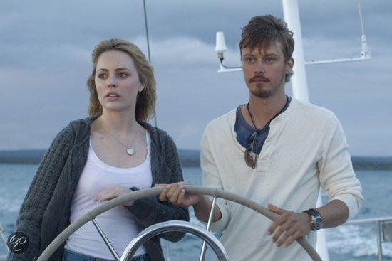 Vrouw met blonde haren en grijze vest aan het roer van een zeilboot, naast een man met baard.