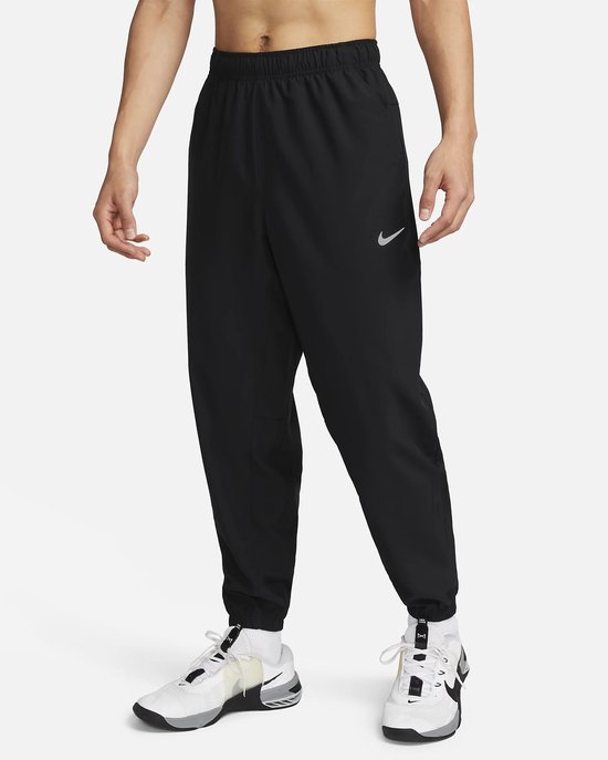Man met zwarte Nike trainingsbroek en witte sportschoenen. Broek heeft een elastische tailleband en geribde enkels. Zilveren Nike logo.