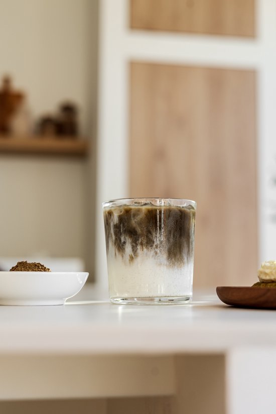 Een glas met ijskoffie met melk, met zichtbare ijsblokjes en swirls van melk en koffie. Op de voorgrond een schaaltje met hojicha poeder.