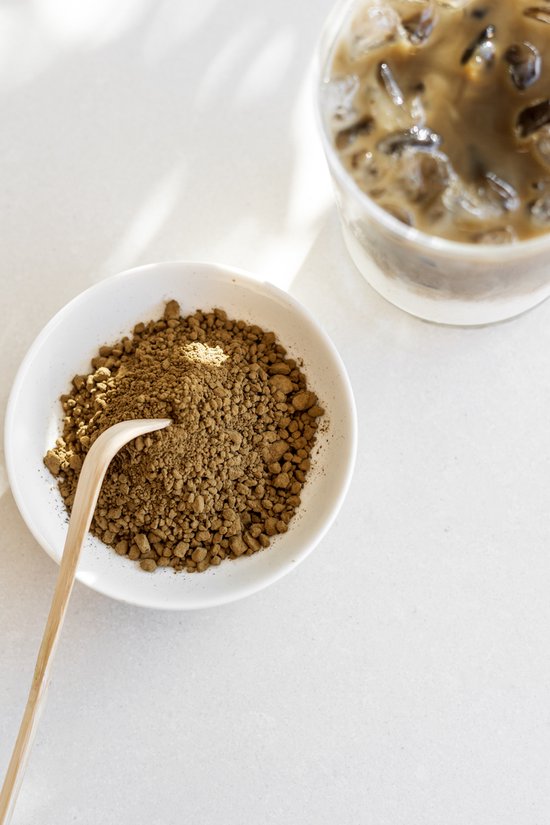 Wit schaaltje met hojicha poeder en een bamboe schepje. Op de achtergrond een glas met ijskoffie.