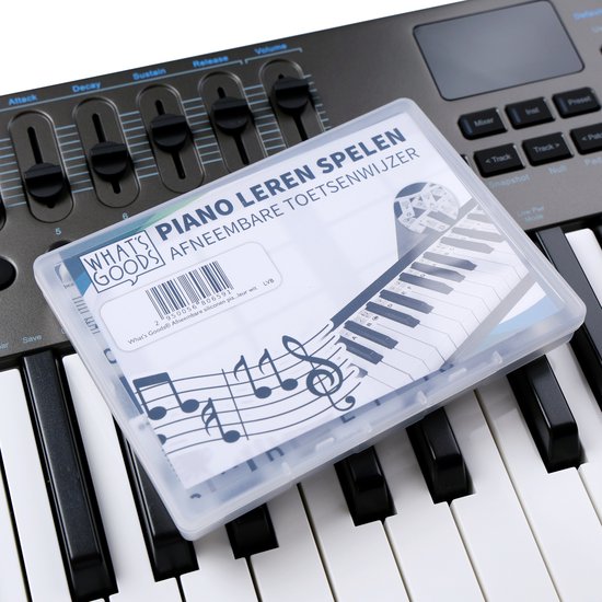 Een doorzichtig doosje met "WHAT'S GOODS PIANO LEREN SPELEN AFNEEMBARE TOETSENWIJZER" op de voorkant, liggend op witte pianotoetsen.