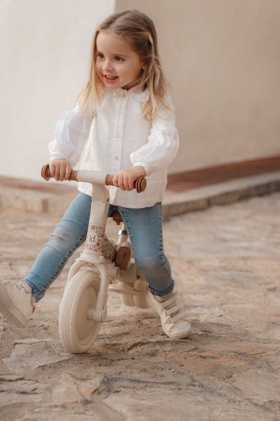 Meisje met blonde staartjes in witte blouse en spijkerbroek rijdt op een crèmekleurige loopfiets met bloemenprint.