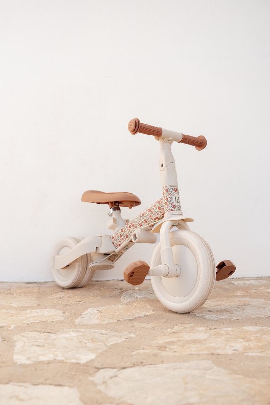 Crèmekleurige loopfiets met bruine zitting en handvatten, op stenen ondergrond. Frame met bloemenprint.