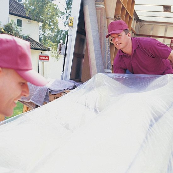 Twee mannen met rode petten kijken naar een opgerolde matras bedekt met plastic folie in een verhuiswagen.