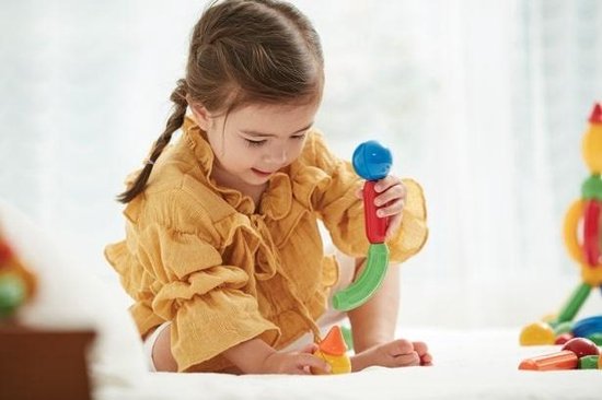 Een jong meisje met een gele blouse speelt met magnetisch speelgoed op een witte ondergrond.